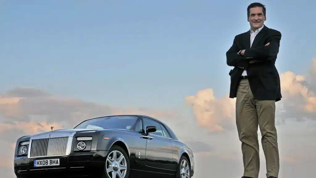 Imagen de Ian Cameron, diseñador de BMW y Rolls-Royce, con los brazos cruzados apoyado en el capó de un automóvil Rolls-Royce. El vehículo está estacionado con un cielo despejado al fondo durante lo que parece ser el atardecer o amanecer, dado el tono del cielo. La placa del vehículo es visible y muestra la inscripción ‘HX08 BHA’.
