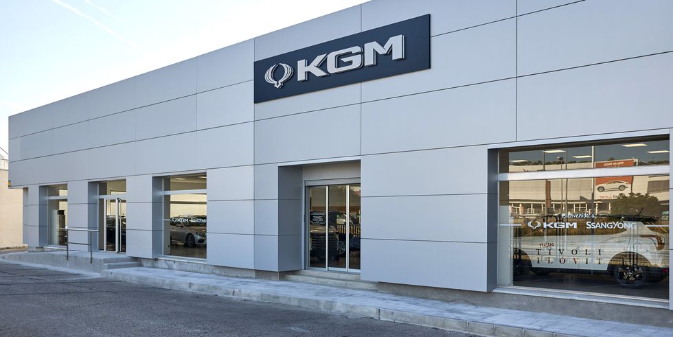 Una imagen del exterior de un edificio moderno con un gran letrero que dice “KGM” en la fachada sobre la entrada. Debajo de este letrero, hay una ventana de vidrio reflectante que muestra otro letrero que dice “KGM SsangYong” con texto adicional debajo que no es claramente legible. El edificio parece ser un concesionario o sala de exposición de automóviles debido a la presencia de coches visibles a través de las ventanas de vidrio.