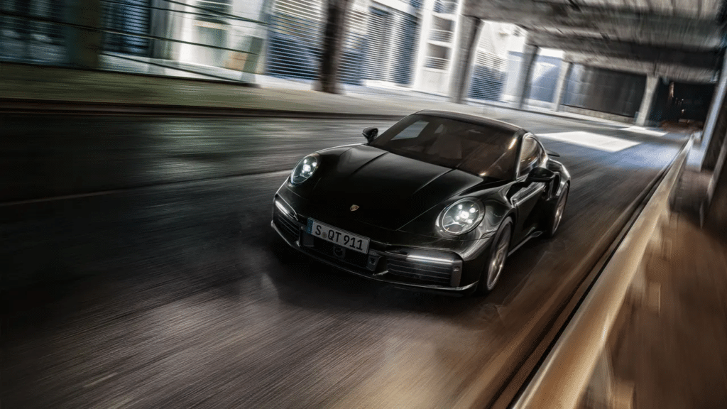 Imagen de un Porsche 911 Turbo S negro en movimiento, capturada con un efecto de desenfoque de movimiento para enfatizar su velocidad y rendimiento dinámico. El coche está centrado en el encuadre, destacando su diseño elegante y estético deportivo.