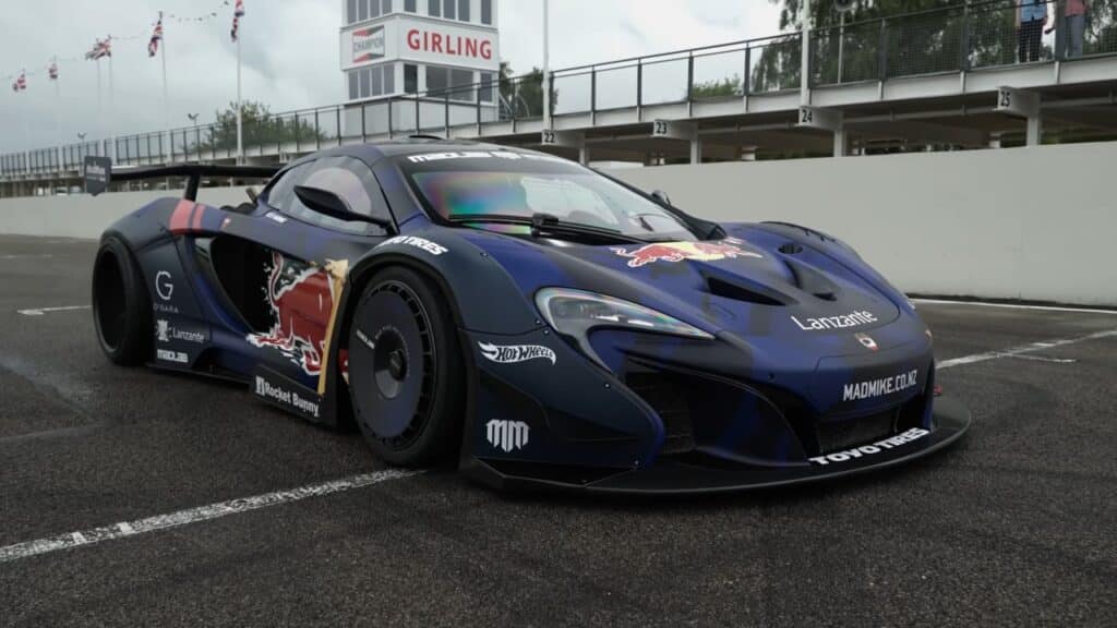 Un coche de carreras McLaren P1, personalizado por Mad Mike, está estacionado en una pista de carreras. El vehículo presenta un esquema de colores oscuros con logotipos prominentes de Red Bull y varios patrocinadores. El coche tiene una postura baja y ancha, con una carrocería aerodinámica y un gran alerón trasero para estabilidad a alta velocidad. Está posicionado frente a un pit lane con el cartel ‘GIRLING’ visible arriba, indicando el contexto de la carrera.