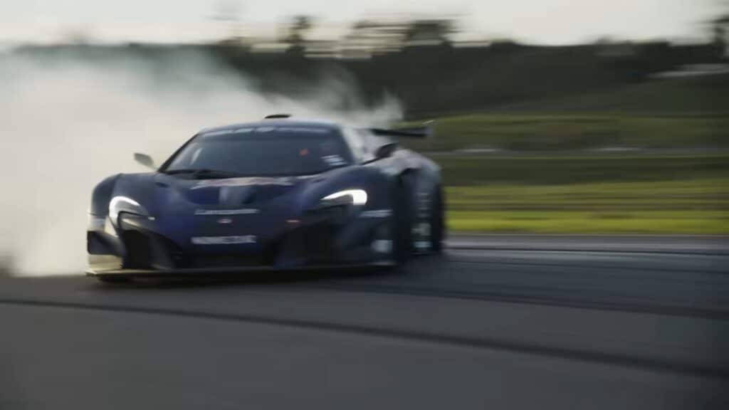 Un coche deportivo McLaren P1, personalizado por Mad Mike, capturado en movimiento en una pista de carreras. El vehículo aparece en una postura dinámica con el fondo borroso, lo que sugiere alta velocidad. La carrocería del coche presenta un trabajo de pintura distintivo con logotipos de marcas y patrocinadores visibles.