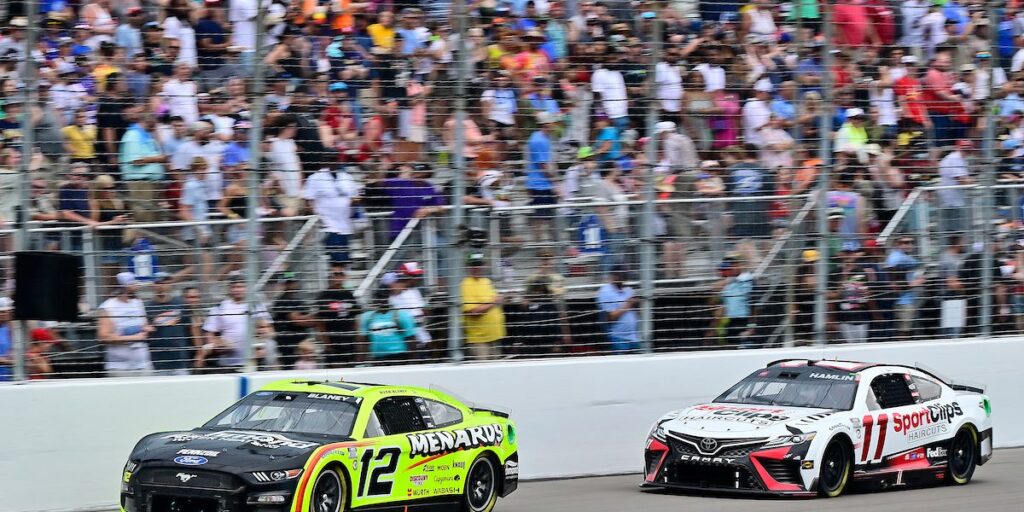 Imagen de dos coches de NASCAR en una pista. Uno es un Ford Mustang negro con el número 12 y logotipos de patrocinadores como ‘Menards’. El otro es un Toyota Camry blanco con el número 11 y patrocinio de ‘Sport Clips’. Los coches están en movimiento sobre una pista de asfalto, con una multitud de espectadores borrosa en el fondo.