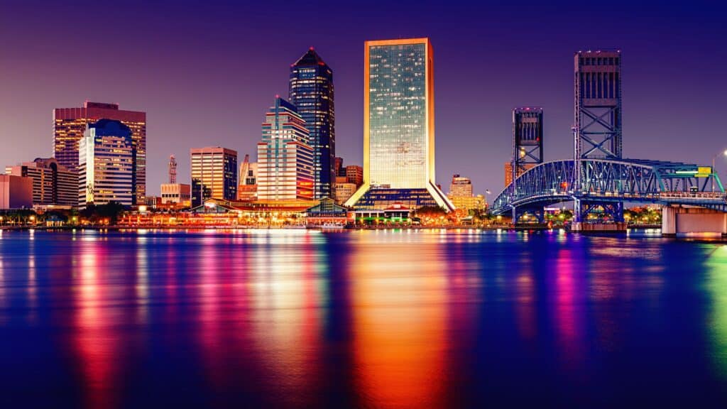 Horizonte de Tampa, Florida, al anochecer, con varios edificios altos, un puente a la derecha y reflejos de las luces en la superficie del agua. El cielo muestra tonos de púrpura y azul. Esta ciudad será la sede de la ZCON de 2024.