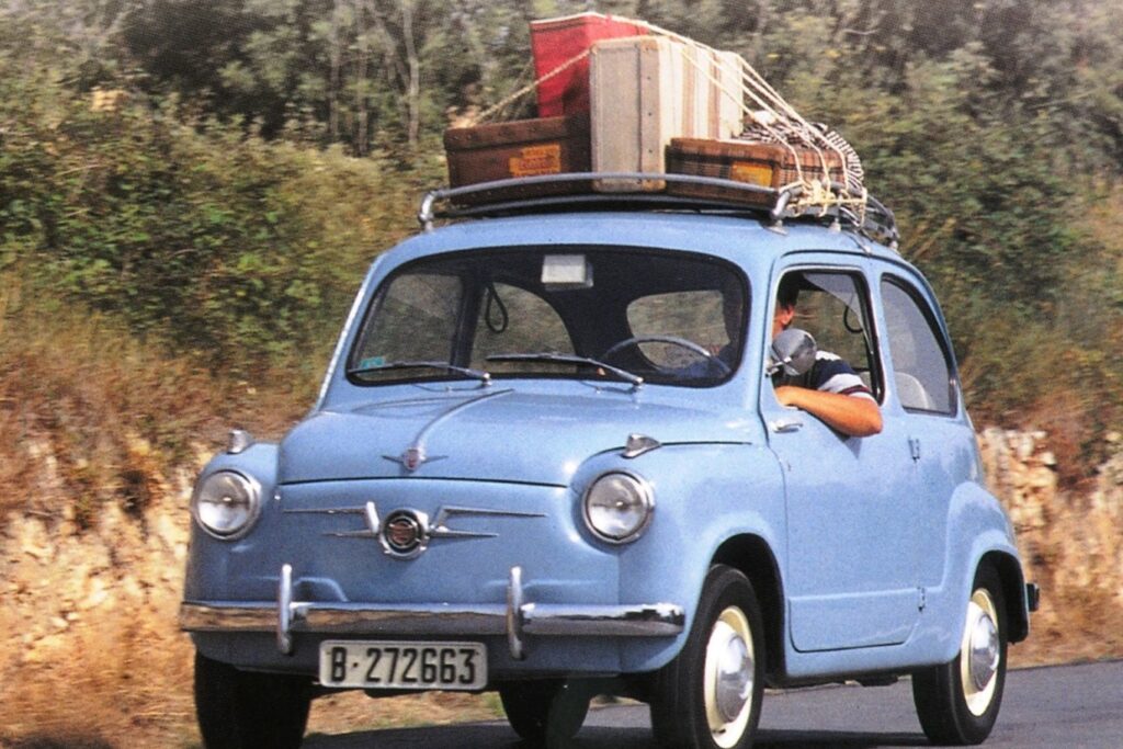 Un Seat 600 azul vintage viaja por una carretera con un paisaje escénico de árboles y vegetación de fondo. El coche, conocido por su tamaño compacto y diseño clásico, lleva equipaje amarrado en el techo, lo que sugiere que los ocupantes están de viaje o de vacaciones.