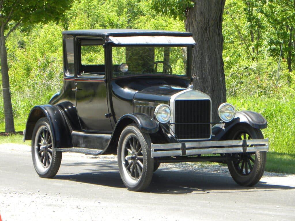 Un automóvil vintage Ford Model T, conocido como “Tin Lizzie”, estacionado sobre una superficie pavimentada con vegetación de fondo. El coche es negro, con una carrocería cuadrada distintiva, una parrilla vertical y grandes faros redondos. Este modelo está bien mantenido o restaurado, reflejando su estatus como una pieza icónica de la historia automotriz.