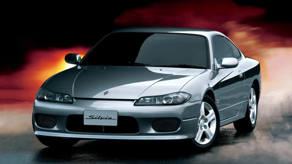 Imagen de un Nissan Silvia S15 de color plateado, un coche deportivo conocido por su rendimiento y popularidad en el tuning y el automovilismo, especialmente en el drifting. La imagen muestra una vista frontal clara, destacando su diseño elegante, faros distintivos y el emblema ‘Silvia’ en la parrilla delantera. El fondo tiene un desenfoque artístico con tonos cálidos que sugieren movimiento o velocidad, enfatizando el aspecto de rendimiento del coche.