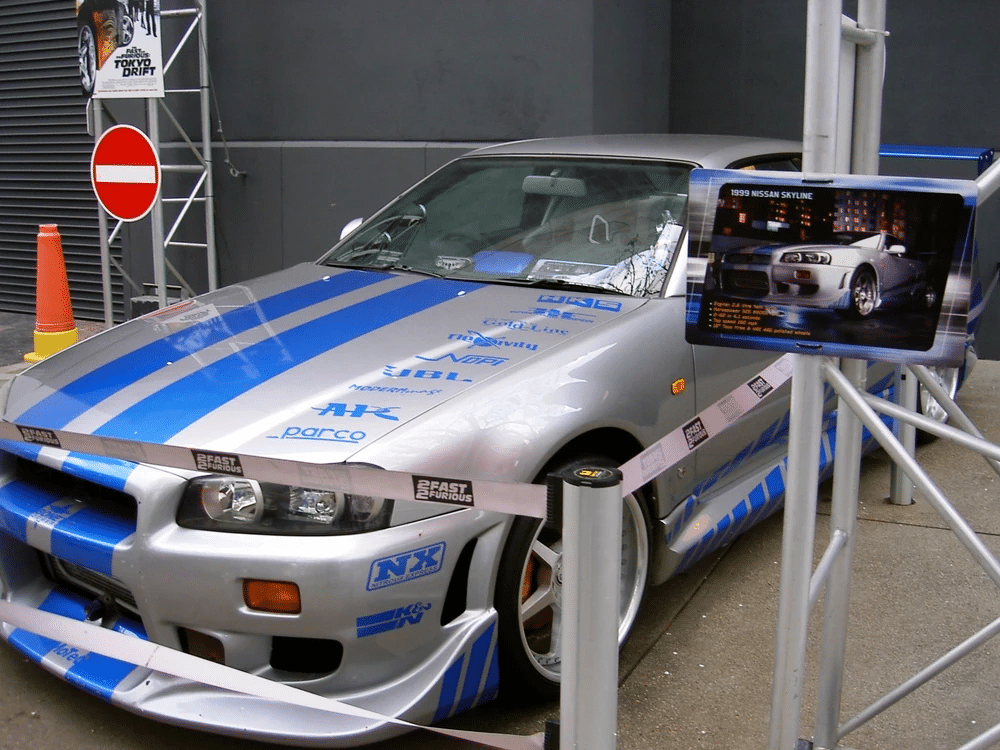Imagen de un automóvil deportivo Nissan Skyline GT-R R34 en colores plateado y azul, estacionado en un área interior. El coche tiene varias marcas y logotipos, incluyendo ‘Nismo’, ‘Sparco’, y ‘Calsonic’. A la izquierda del automóvil hay un cono de tráfico naranja y una torre metálica, y a la derecha hay una pantalla informativa montada en un soporte. El coche está rodeado por una cinta que dice ‘2Fast 2Furious’.