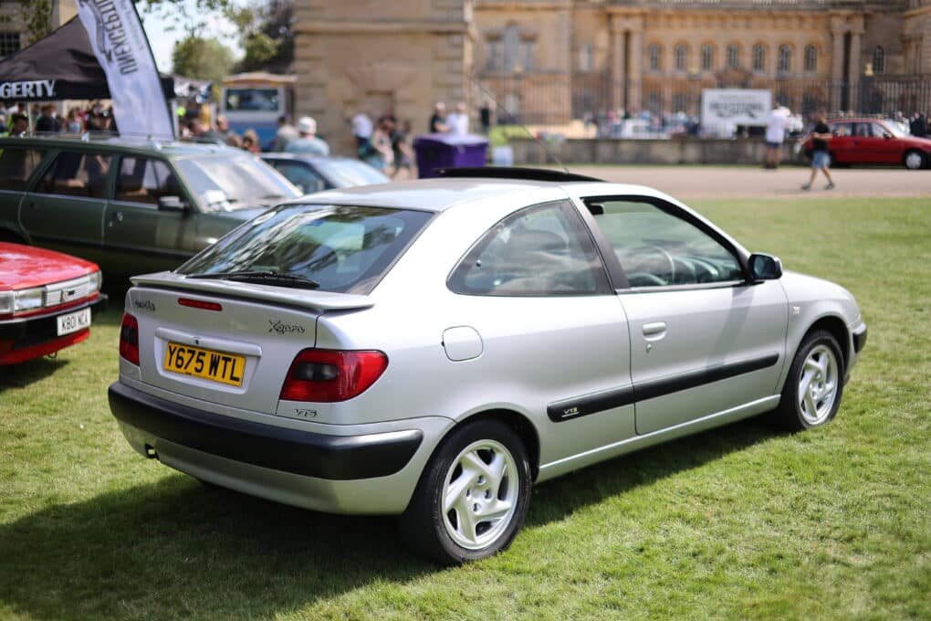 Un Citroën Xsara plateado de 2001 está estacionado sobre el césped durante el Festival of the Unexceptional. En el fondo, se ve un edificio y otros coches antiguos. El coche tiene una carrocería tipo hatchback y la matrícula es Y67 MTL.