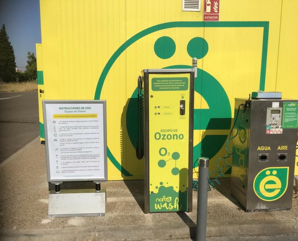 Imagen de una sección de la gasolinera Nafte con una pared amarilla brillante. A la izquierda, hay un soporte blanco con instrucciones en español, y a su derecha, un equipo de ozono etiquetado ‘Equipo de Ozono’ en colores verde y amarillo. Junto a este, hay una máquina verde y gris etiquetada ‘AGUA AIRE’ para servicios de agua y aire. El suelo es de hormigón seco y la zona está bien iluminada, sugiriendo que la foto fue tomada durante el día. Esta área fue fotografiada antes de que derribaran la pared debido a un presunto robo.
