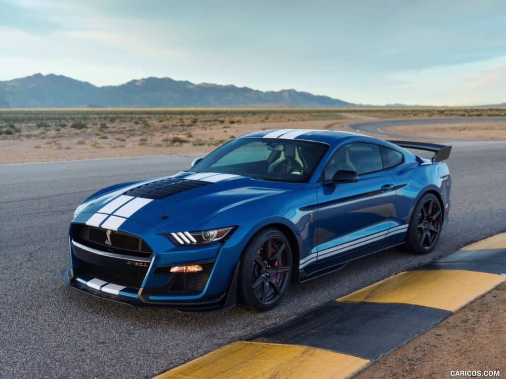 Imagen de un Ford Mustang GT500 azul con franjas blancas de carreras, estacionado sobre una superficie de asfalto. El coche está posicionado en un ángulo que muestra su perfil frontal y lateral, con un fondo de paisaje desértico bajo un cielo despejado. Este modelo parece ser de los años 2020 a 2022.