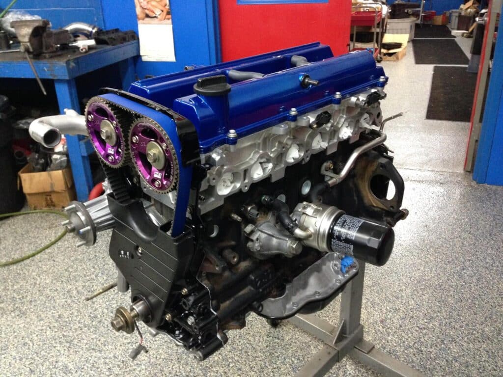 Imagen de un motor 2JZ de Toyota. El motor está montado en un soporte en un taller o garaje, posiblemente para mantenimiento o modificación. Se observan algunas piezas de posventa instaladas, como una polea de leva en colores púrpura y azul, y posiblemente otras mejoras de rendimiento.
