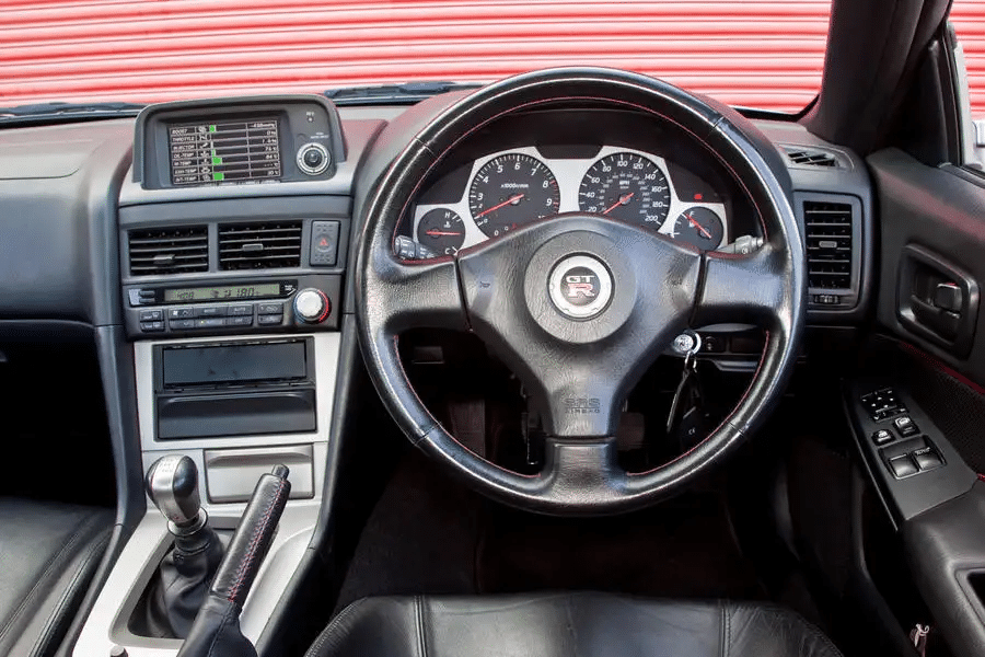 Interior de un Nissan Skyline GT-R R34, centrado en el lado del conductor. Se observa un volante con un emblema en el centro. A la izquierda del volante está el tablero del vehículo, que incluye varios controles y un sistema de audio. La palanca de cambios es visible en la consola central, y hay líneas horizontales rojas en el fondo, posiblemente de una puerta de garaje o pared detrás del coche.