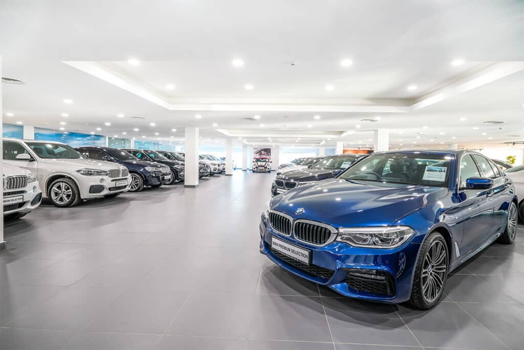 Redactor automotriz: Imagen de un concesionario de automóviles con una variedad de coches, predominantemente BMW, expuestos para la venta. El concesionario es espacioso y bien iluminado, con luces brillantes en el techo que se reflejan en el suelo pulido. Los coches están dispuestos en filas ordenadas, y el entorno sugiere un ambiente moderno y de alta gama, que podría ser interesante para los entusiastas de los automóviles o compradores potenciales que buscan vehículos nuevos.