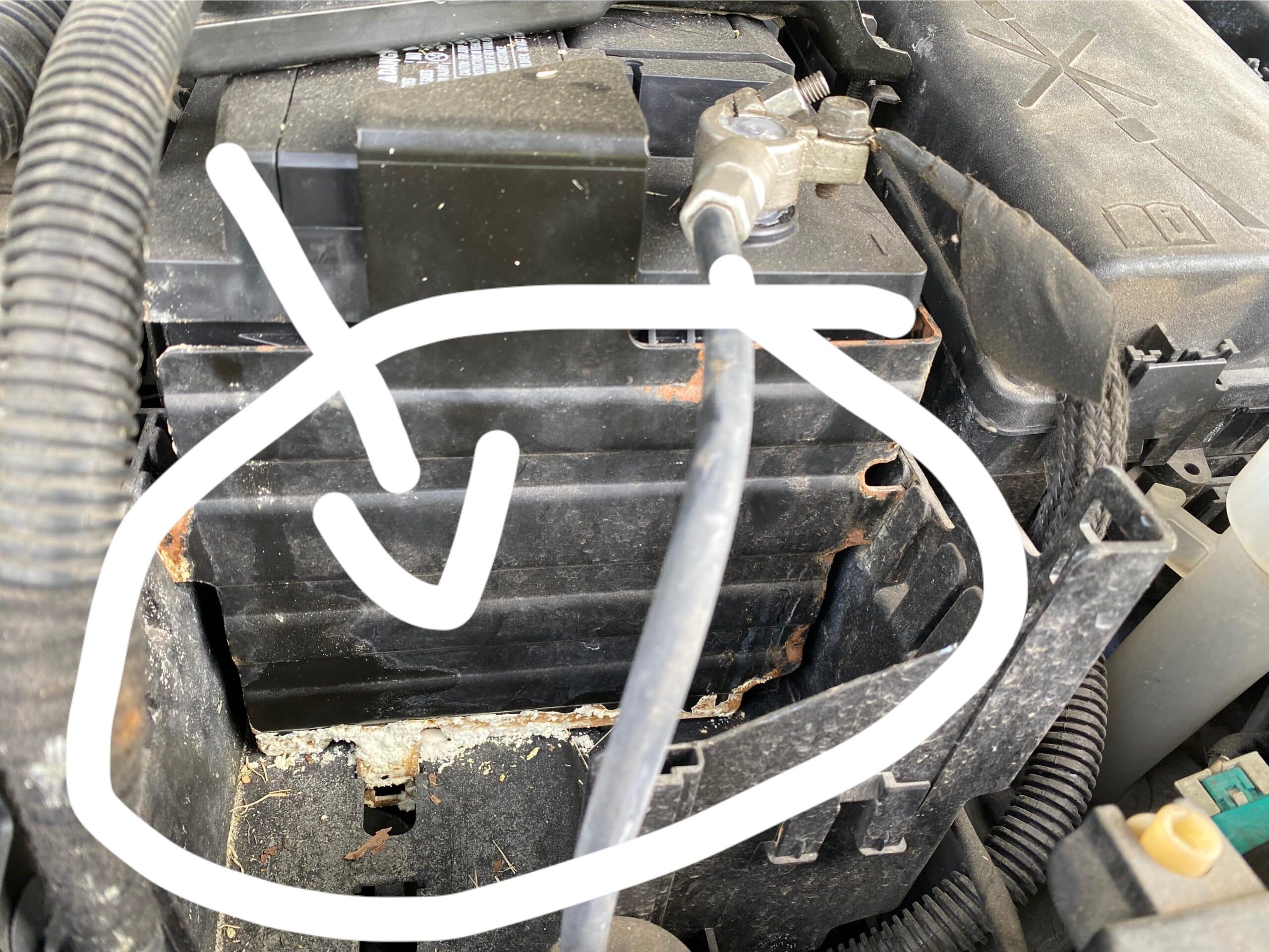 La imagen muestra una vista de cerca del compartimento del motor de un vehículo, enfocándose en un componente específico que parece ser la carcasa del filtro de aire. Este componente presenta daños significativos, con corrosión visible y degradación del material, probablemente causada por una fuga de la batería. Esto es relevante ya que indica posibles problemas de mantenimiento del vehículo que podrían afectar a su rendimiento.