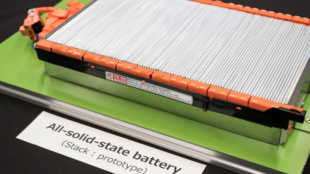 Un prototipo de batería de estado sólido colocado sobre una superficie verde. La batería es rectangular, con múltiples módulos metálicos grises apilados uno al lado del otro, encerrados en un marco naranja. Hay una etiqueta con un símbolo de advertencia y texto en el borde superior de la batería, aunque el texto no es legible. Delante de la batería, hay una etiqueta blanca que dice “All-solid-state battery (Stack - prototype)”