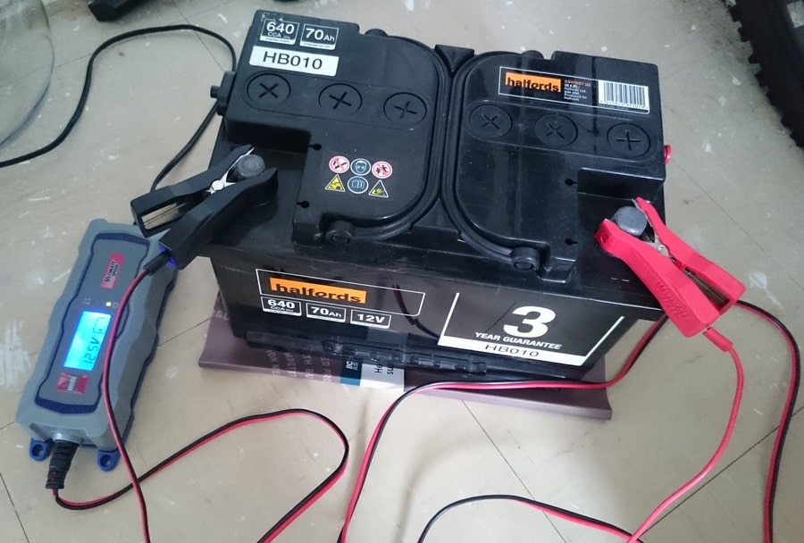 Batería de coche en el suelo con un cargador de batería conectado, mostrando claramente los terminales positivo y negativo con los signos más (+) y menos (-), y las pinzas del cargador sujetas a los terminales correspondientes.