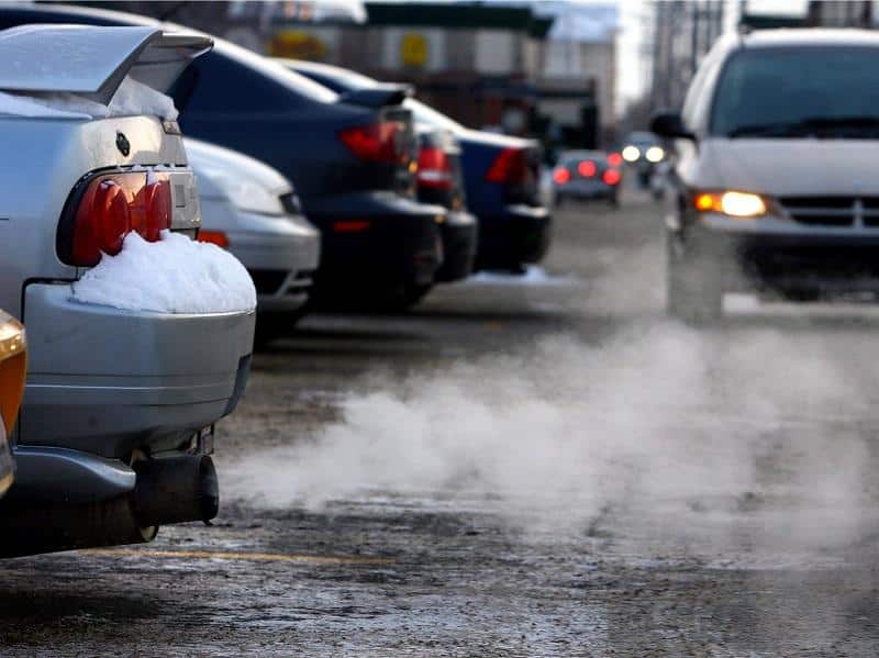 Imagen de la parte trasera de un coche con humo visible saliendo del tubo de escape. El coche está estacionado y parece estar a ralentí, posiblemente después de un cambio de aceite. En el fondo, se ven otros coches, lo que sugiere que podría ser una escena de tráfico o un área de estacionamiento.