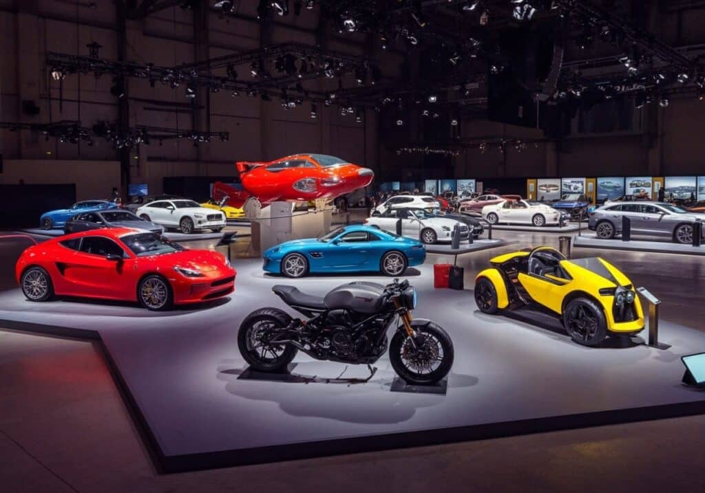 Redactor automotriz: Imagen de una exhibición de vehículos en un salón del automóvil. En primer plano, hay un coche deportivo rojo, detrás de él un coche deportivo azul, y varios otros coches de diferentes modelos y colores en el fondo. Suspendido del techo, hay un avión rojo. En el centro de la imagen hay una motocicleta negra, y a su derecha un vehículo amarillo de tres ruedas. El entorno parece ser un pabellón de exposiciones con focos que iluminan cada vehículo.