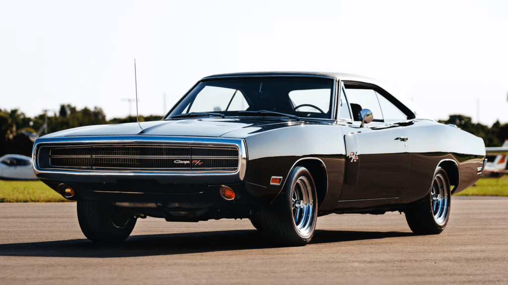 Imagen de un Dodge Charger R/T de 1970, conocido como el coche de Toretto en The Fast and the Furious. El coche está estacionado en lo que parece ser una pista de aterrizaje, con el sol bajo en el horizonte, dando a la foto un tono cálido. El coche es negro con detalles cromados y tiene características distintivas de un muscle car, como un capó largo, dos puertas y suspensión trasera elevada.