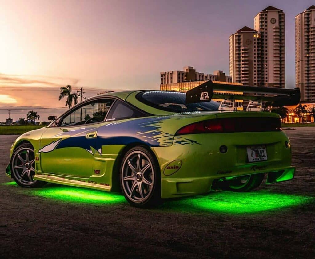 El primer coche de Brian en Fast and Furious: Imagen del Eclipse GSX de color verde con franjas y detalles en azul, estacionado al aire libre con edificios al fondo durante el amanecer o atardecer. El coche tiene piezas personalizadas, incluyendo un gran alerón trasero y luces de neón verdes que iluminan el suelo.