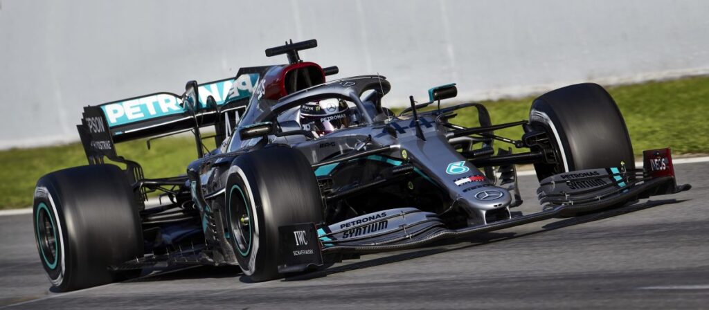 La imagen muestra un coche de carreras de Fórmula 1 Mercedes-Benz Petronas en una pista. El coche es predominantemente negro con acentos en color verde azulado y verde, y presenta el logotipo de Petronas de manera prominente. El vehículo está equipado con neumáticos lisos de competición y una carrocería aerodinámica compleja, incluyendo alerones delanteros y traseros diseñados para estabilidad a alta velocidad y en curvas. It's hammer time.