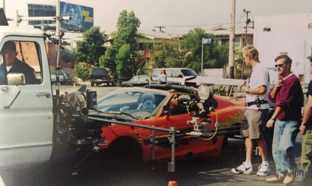 Momento detrás de cámaras del rodaje de ‘The Fast and the Furious’. Se muestra un Toyota Supra naranja con un equipo de cámara adjunto a su lado. Varias personas están alrededor del coche, una operando la cámara y otra observando. El escenario parece ser una calle urbana con otros vehículos y edificios al fondo.