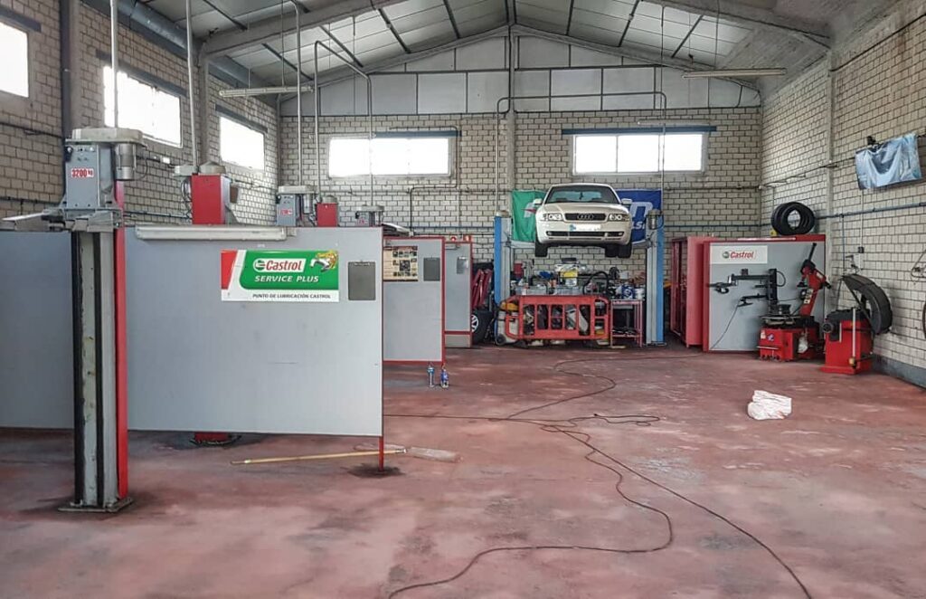 Donde cambiar el aceite del coche uno mismo en Madrid: Imagen del interior de un centro de servicio de vehículos llamado GT BOX CENTER. Hay un coche elevado en un elevador para mantenimiento o reparación, y varias piezas de equipo y herramientas visibles alrededor del taller, como una máquina para cambiar neumáticos, bancos de trabajo y unidades de almacenamiento. La presencia del logotipo de la marca Castrol sugiere que podrían usar o suministrar productos Castrol.