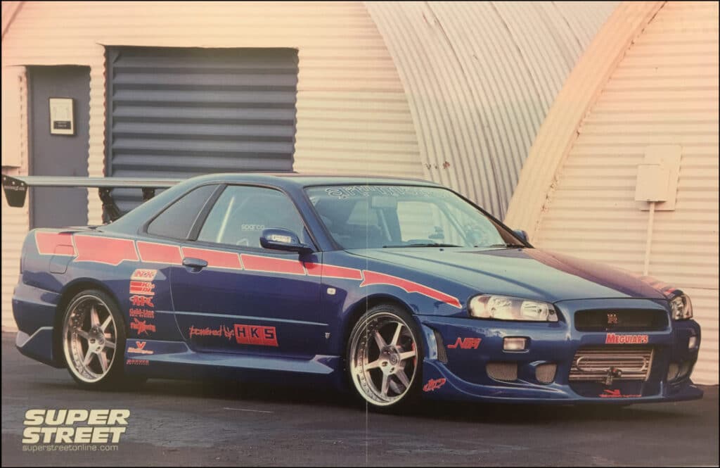 Nissan Skyline azul con un gran alerón trasero y varias calcomanías y logotipos de patrocinadores en su carrocería. Este coche, propiedad de Craig Lieberman, fue modificado posteriormente para la película ‘2 Fast 2 Furious’. La imagen está tomada en un área industrial o un garaje.