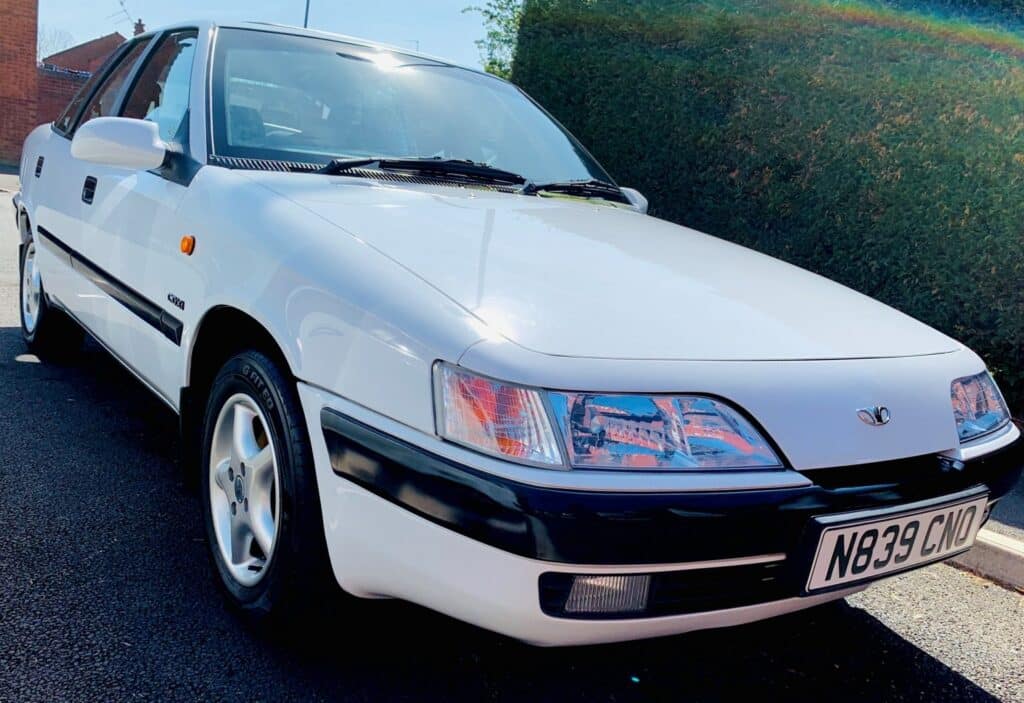 Un Daewoo Aranos blanco, también conocido como Daewoo Espero, está estacionado al aire libre bajo un cielo despejado durante el Festival of the Unexceptional. El vehículo está orientado hacia la izquierda, mostrando su parte frontal derecha. El coche presenta un diseño distintivo de los años 90 con faros emergentes y un borde negro en la parte inferior de la carrocería. La matrícula es visible.