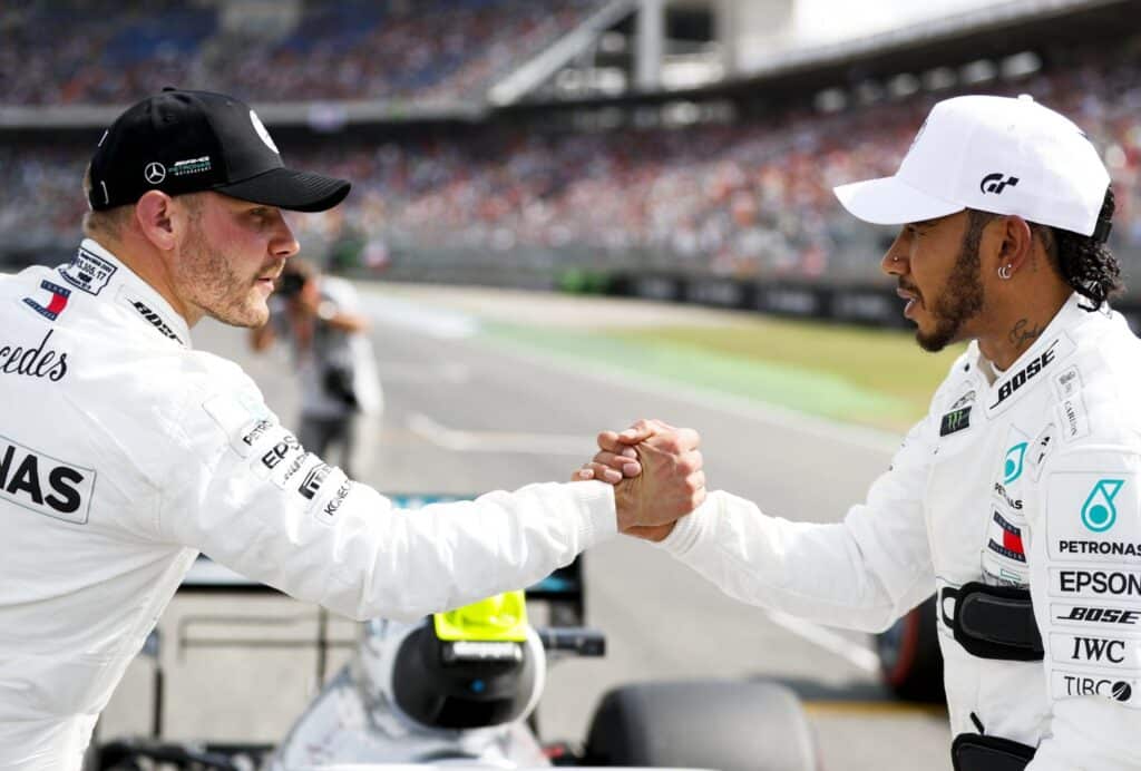 Lewis Hamilton y Valtteri Bottas, ambos con monos de carreras blancos con múltiples logotipos de patrocinadores como Mercedes y PETRONAS, se dan la mano. Este gesto podría indicar deportividad o una celebración entre los dos pilotos.