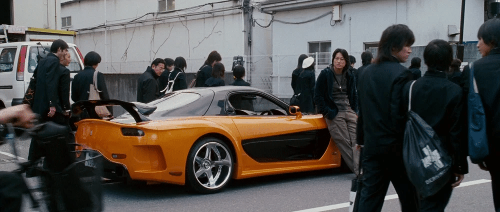 Imagen de un Mazda RX-7 Veilside con kit Fortune, el coche de Han en ‘Fast and Furious’. El coche es de color naranja brillante y está estacionado en la calle. Han está apoyado en el coche. Hay varias personas alrededor del coche.