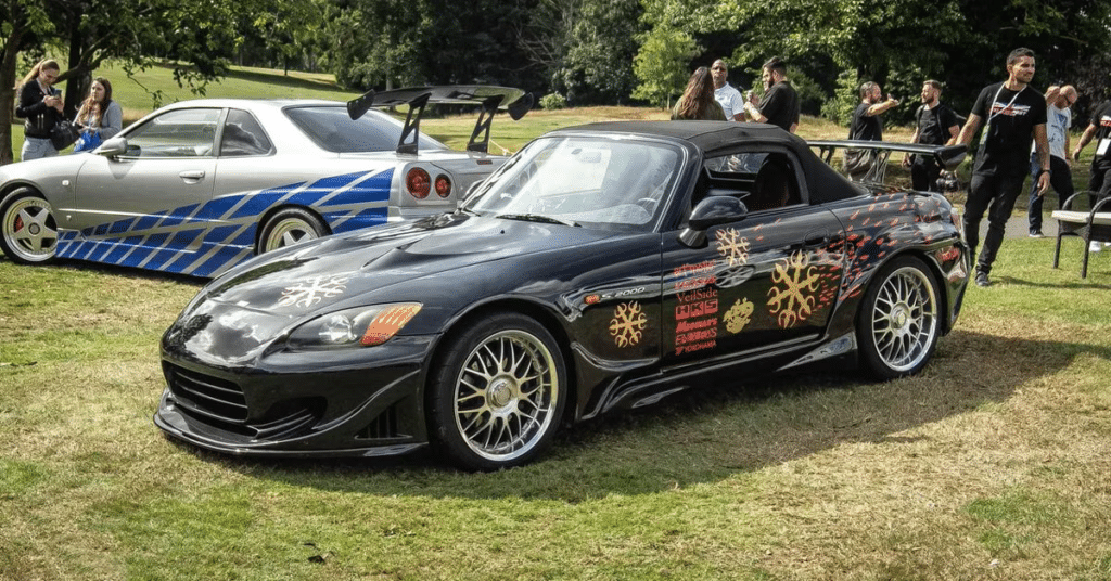 Honda S2000 de Fast and Furious 1, negro con un diseño personalizado de salpicaduras blancas y rojas en el lateral. El coche está estacionado sobre césped en lo que parece ser una exhibición de autos, con otros coches personalizados y espectadores en el fondo.