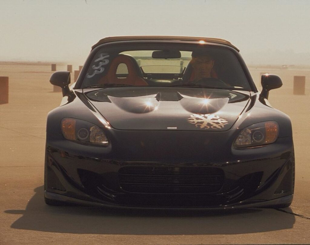 Vista frontal del Honda S2000 de Fast and Furious 1, negro con gráficos personalizados, con los faros encendidos. Una persona es visible a través del parabrisas, sentada en el asiento del conductor. El coche está estacionado en un lote vacío con un cielo despejado de fondo.