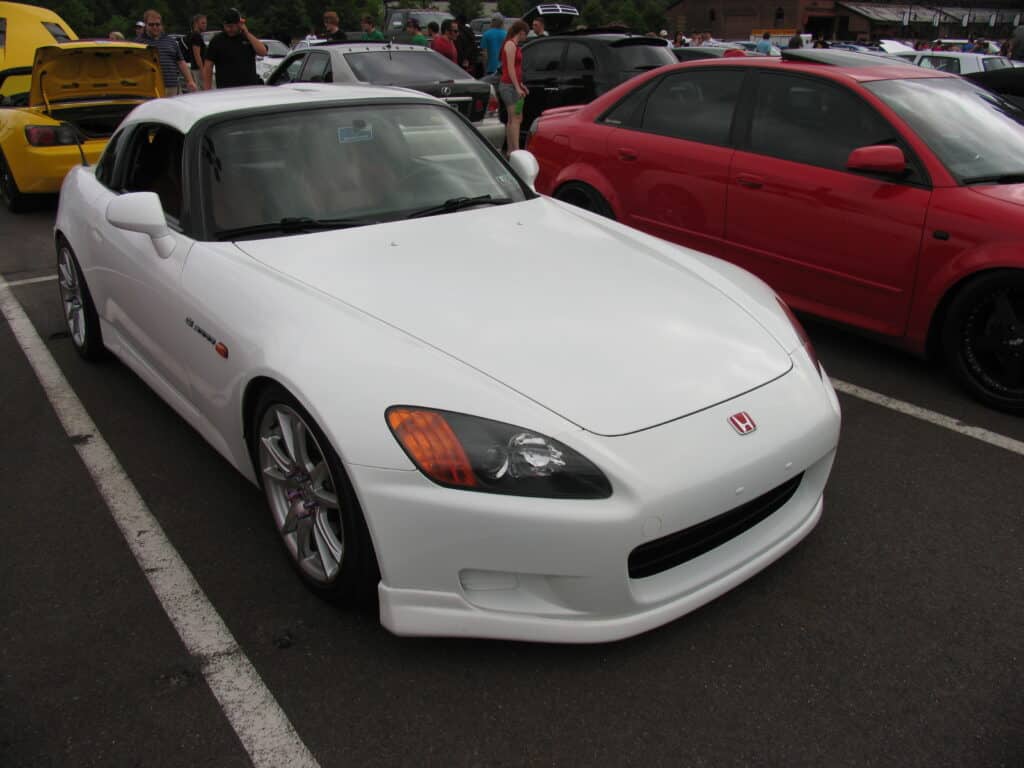 Un coche deportivo Honda S2000 blanco está estacionado en un aparcamiento junto a otros vehículos. El coche presenta un diseño elegante con un capó largo y una parte trasera corta, típico de los roadsters. Tiene el emblema de Honda en la parte delantera y parece estar en buen estado. En el fondo se pueden ver varios otros coches, incluyendo un vehículo amarillo y uno rojo, lo que indica que podría ser un encuentro de coches o un evento similar.