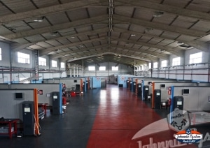 Donde cambiar el aceite del coche uno mismo en Madrid: Imagen del interior de un gran espacio industrial o taller Johnnie Pyston. Hay varias máquinas grandes, posiblemente para fines de fabricación o ingeniería, alineadas a ambos lados del espacio. El techo es alto con cerchas visibles y tragaluces que permiten la entrada de luz natural. El suelo parece ser de hormigón y hay un pasillo central para caminar o mover objetos.