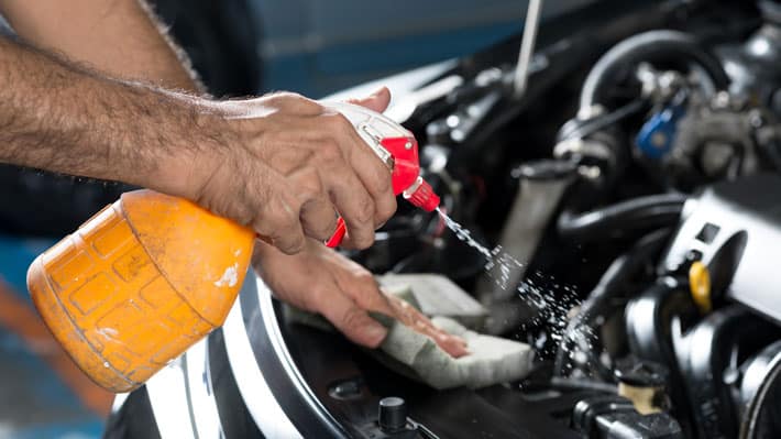 Las manos de una persona usando una botella de spray para aplicar un líquido sobre un paño en una parte del motor de un vehículo. Esta acción sugiere que se está realizando mantenimiento o limpieza en el motor del vehículo.