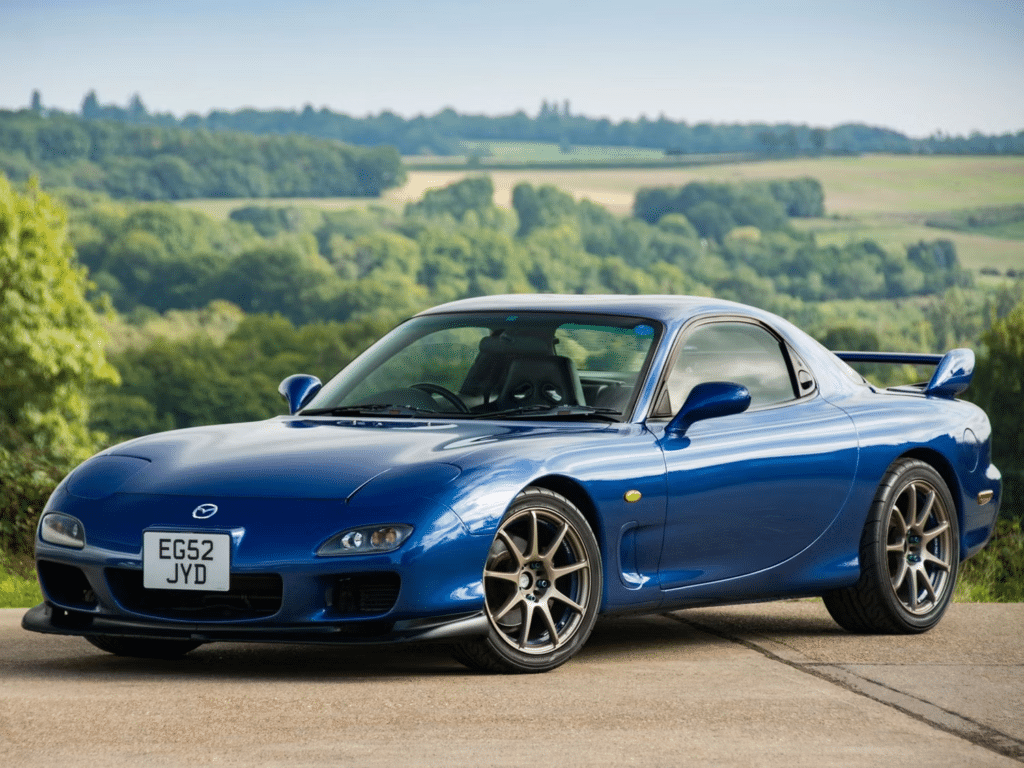 Imagen de un Mazda RX-7 de tercera generación de color azul estacionado al borde de una carretera, con un paisaje natural de fondo. El coche tiene un diseño aerodinámico con dos puertas y faros escamoteables cerrados. La matrícula visible muestra ‘EG52 JYD’.