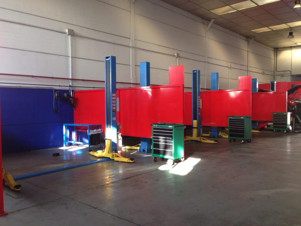 Donde cambiar el aceite del coche uno mismo en Madrid: Imagen de un taller de mantenimiento de vehículos, probablemente Mecánica Box Parla. Hay varios elevadores rojos para coches, uno de los cuales está en uso con un coche elevado y el capó abierto. El taller parece estar bien equipado con herramientas y cajas de herramientas móviles, lo que sugiere un entorno profesional para la reparación y mantenimiento de vehículos.