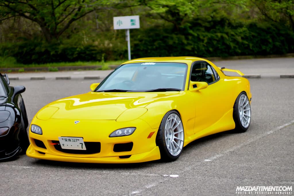 Un Mazda RX-7 FD3S amarillo brillante estacionado en un entorno al aire libre con árboles y vegetación en el fondo. El vehículo está en el lado derecho del marco, con una vista parcial de otro coche a su izquierda. El Mazda RX-7 FD3S es notable por su diseño elegante, faros emergentes y es un modelo popular entre los entusiastas del automóvil por su exclusivo motor rotativo.