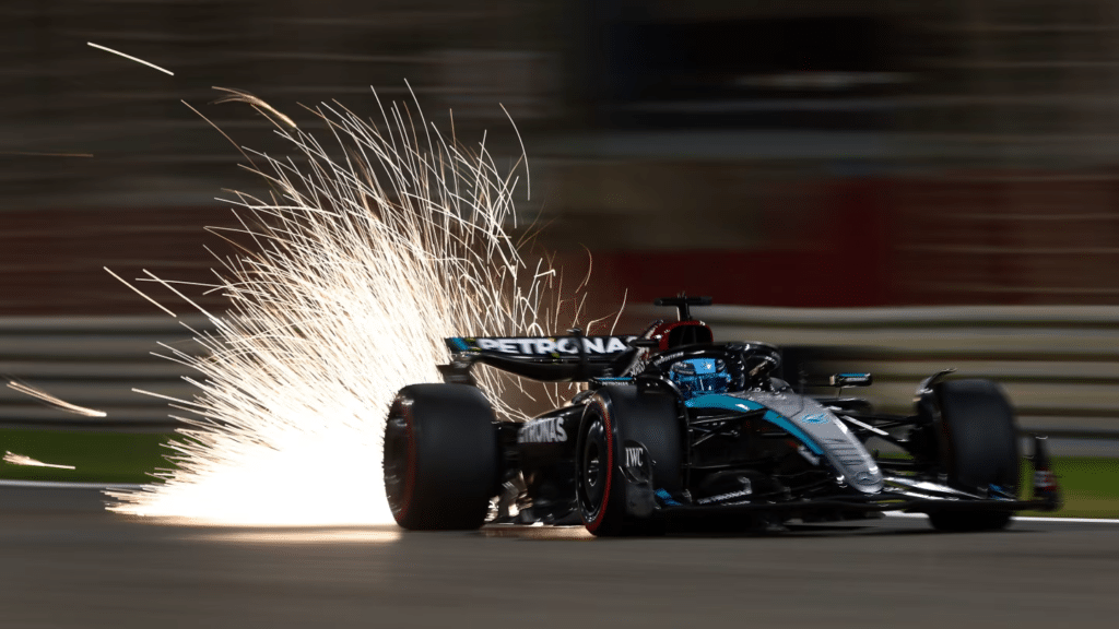 Un coche de Fórmula 1 de Mercedes-Benz en una pista a alta velocidad durante el atardecer. Vuelan chispas dramáticamente desde la parte trasera del coche al hacer contacto con la pista, destacando su velocidad y potencia. El coche es mayoritariamente negro con detalles en verde azulado y blanco, y lleva el logotipo de Mercedes. El fondo está borroso debido al movimiento, enfatizando la velocidad del coche.