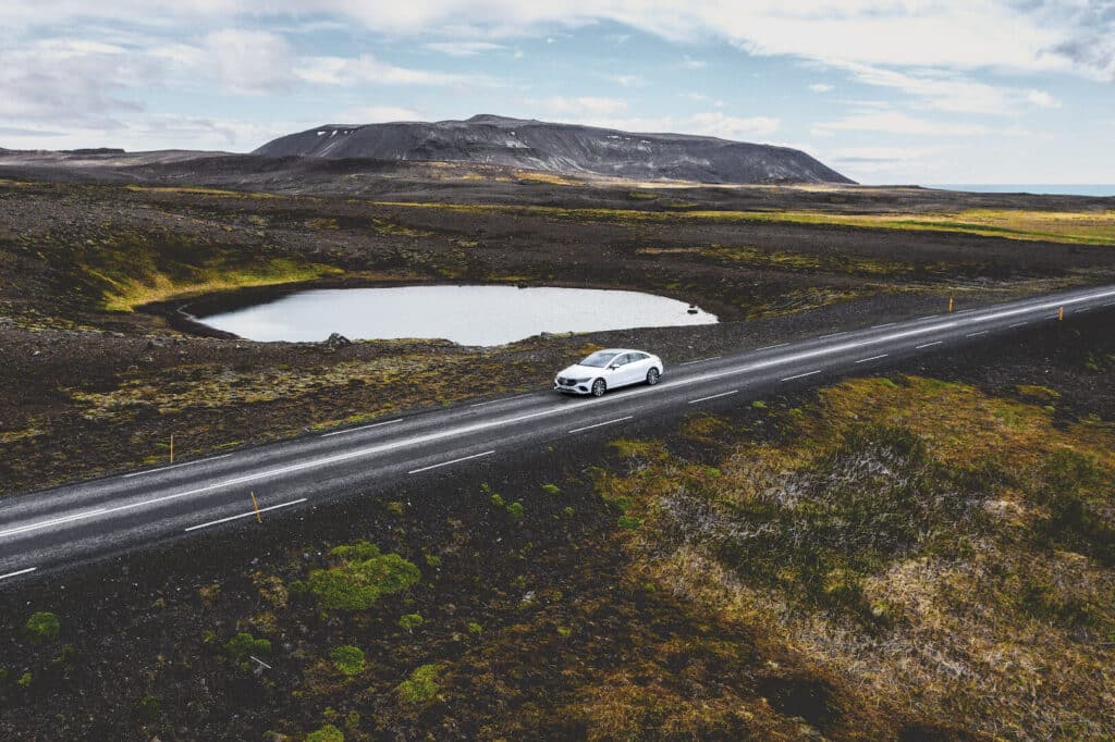 Un coche de Mercedes-Benz blanco estacionado al lado de una carretera que atraviesa un paisaje agreste con vegetación escasa. El terreno es mayoritariamente plano con un pequeño cuerpo de agua cercano y colinas en la distancia bajo un cielo nublado. La imagen sugiere una armonía entre la tecnología y la naturaleza, destacando el enfoque en las energías verdes y la sostenibilidad.