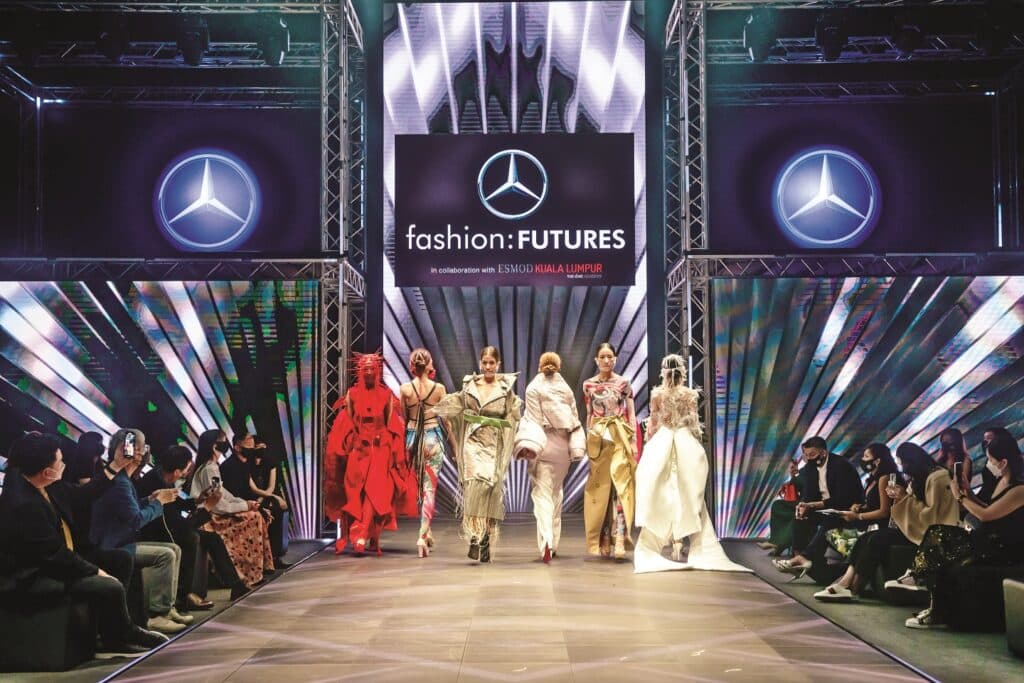 Un evento de la Mercedes-Benz Fashion Week. Hay una pasarela con modelos desfilando, mostrando varios conjuntos. El público está sentado a ambos lados de la pasarela, observando a los modelos. Al final de la pasarela, hay una gran pantalla con el logotipo de Mercedes-Benz y el texto “fashion:FUTURES”. El entorno sugiere que es un evento que destaca las tendencias y diseños de moda del futuro.
