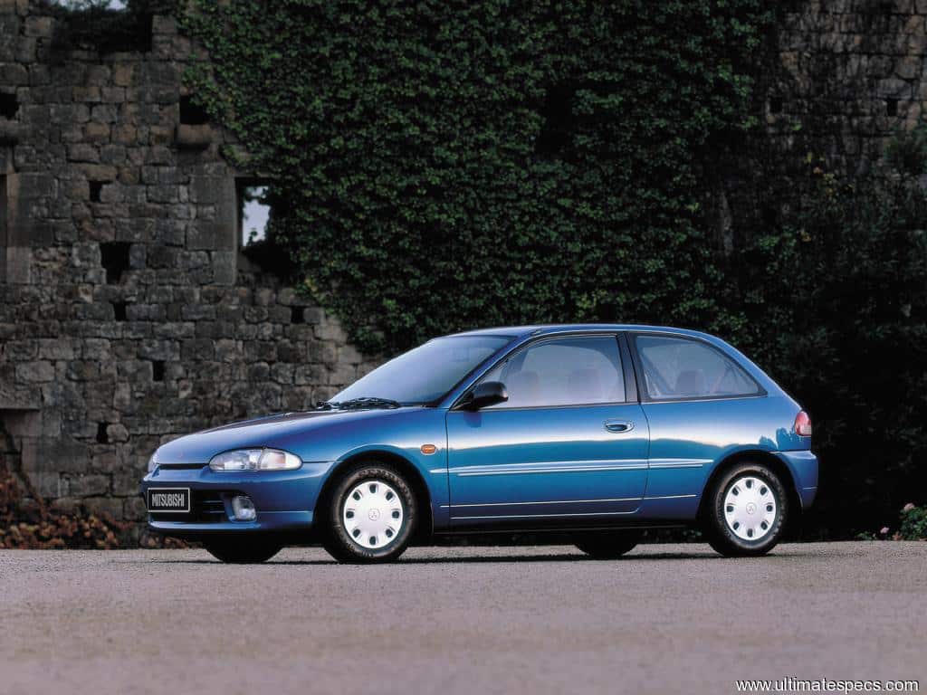 Un Mitsubishi Colt azul está estacionado frente a un muro de piedra, posiblemente parte de una estructura o edificio antiguo. El coche está posicionado en el lado izquierdo del encuadre, permitiendo una vista clara de su perfil. Tiene tapacubos blancos y la matrícula es visible. Esta foto no fue tomada en el Festival of the Unexceptional.