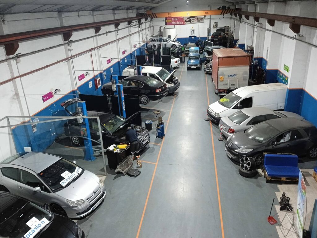 Interior de Motor Home, un taller de alquiler de boxes donde cambiar el aceite del coche uno mismo en Zaragoza. Se observan varios coches en diferentes estados de reparación, algunos elevados en elevadores y otros en el suelo. El espacio es amplio, con techo alto y equipado con herramientas profesionales.