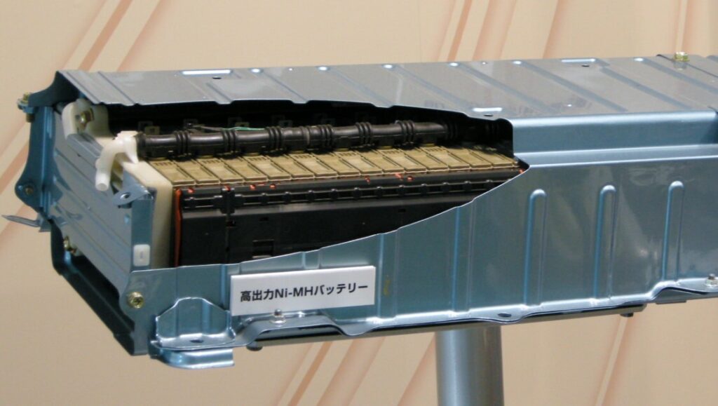 Un módulo de batería de hidruro de níquel-metal (NiMH) para vehículos híbridos. El módulo está parcialmente encerrado en una carcasa metálica con aletas de enfriamiento visibles, indicando su integración en un sistema de gestión térmica. Hay texto en japonés en el lateral de la batería, probablemente con información sobre el modelo o especificaciones.