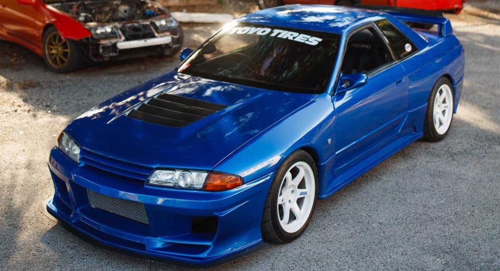 Un Nissan Skyline GT-R R32 azul con un kit de carrocería personalizado, que incluye un capó ventilado, un parachoques delantero extendido y faldones laterales. El vehículo está estacionado sobre una superficie de asfalto con otro coche parcialmente visible en el fondo. Tiene llantas blancas de posventa y una pancarta en el parabrisas que dice ‘TOYO TIRES’. El diseño elegante y las modificaciones del coche sugieren que está construido para conducción de alto rendimiento.