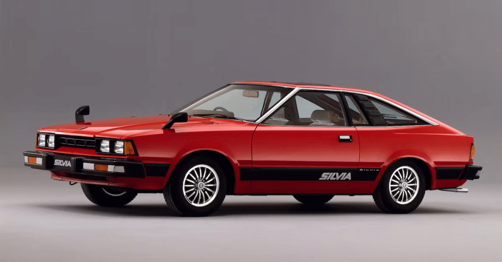 Imagen de un Nissan Silvia S110 de color rojo, mostrando su diseño clásico con faros escamoteables, una carrocería de dos puertas y detalles distintivos en negro alrededor de las ventanas y en la parte inferior del vehículo. Las ruedas tienen un diseño de múltiples radios. La imagen captura el coche desde un ángulo lateral, destacando su perfil deportivo y elegante.