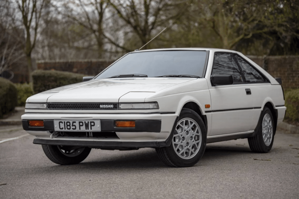 Imagen de un Nissan Silvia S12 de color blanco, estacionado al aire libre. El vehículo tiene una carrocería coupé de dos puertas, faros escamoteables y un diseño distintivo de los años 80. Las ruedas son de aleación y la imagen muestra el coche desde un ángulo que permite ver claramente su perfil frontal y lateral.