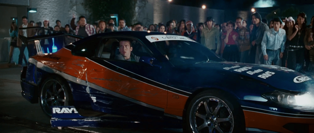 Imagen de un Nissan Silvia s15 en color azul y naranja, estacionado con su parte frontal ligeramente orientada a la izquierda. Hay varias personas de pie en el fondo, detrás de barreras, observando el coche. Este es el coche de Sean en Fast and Furious: Tokyo Drift, conocido por ser destruido en la película.