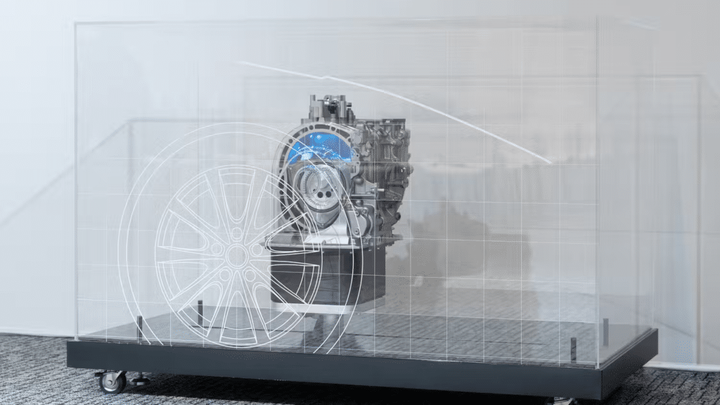 Imagen de un motor rotativo de Mazda exhibido en una plataforma transparente con ruedas. Se observa la estructura interna del motor, incluyendo el rotor y otras partes mecánicas, así como una rueda de automóvil adjunta a un extremo del motor. La presentación destaca la ingeniería y el diseño compacto del motor rotativo.